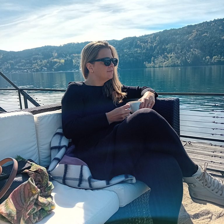 Frau sitzt entspannt am Ufer eines Sees, trägt Sonnenbrille und hält eine Kaffeetasse in der Hand.