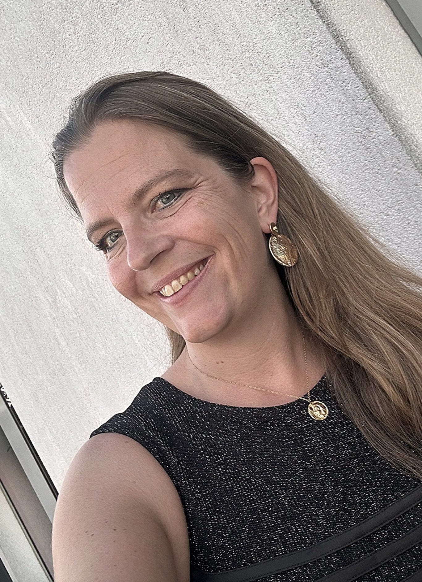 Eine lächelnde Frau mit langen, dunkelblonden Haaren und Ohrringen. Sie trägt ein schwarzes Oberteil. Das ist die Autorin.