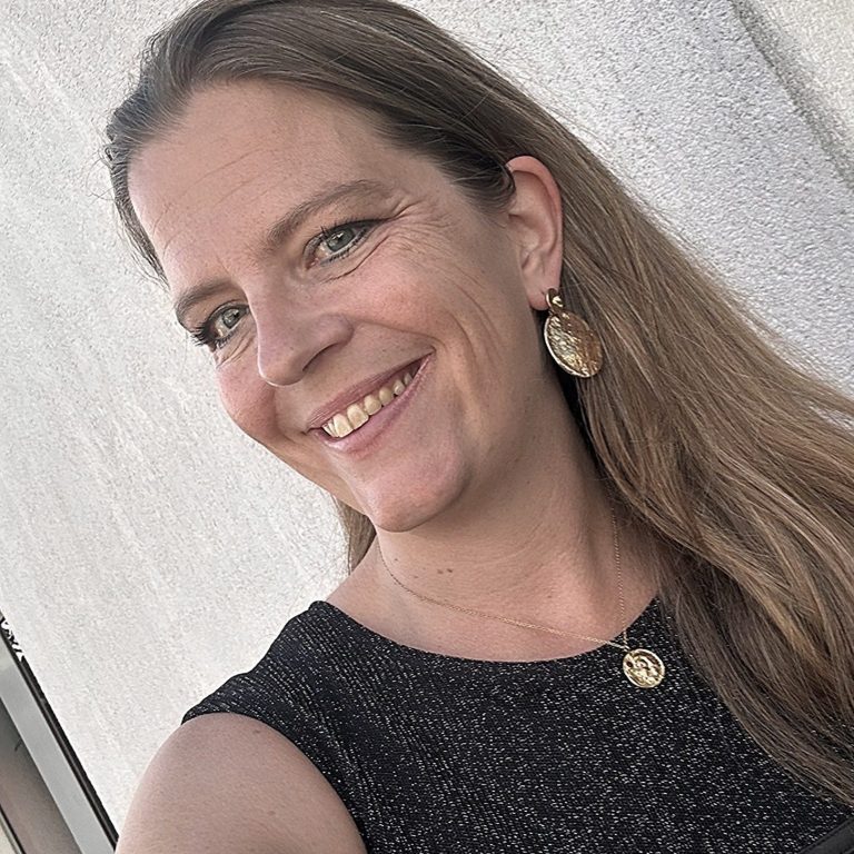 Frau mit langen, blonden Haaren und Ohrringen, lächelt vor einer hellen Wand.