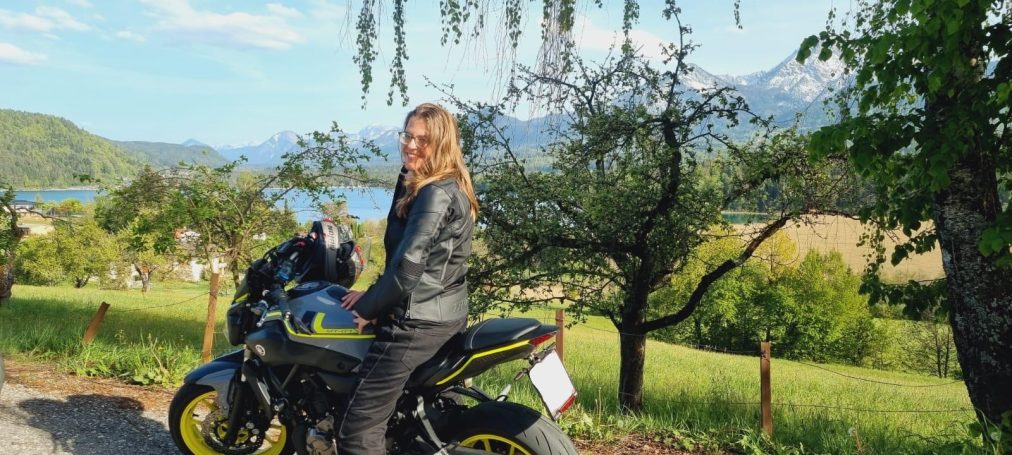 Person mit längeren Haaren sitzt auf einem Motorrad, umgeben von Bäumen und Landschaft.