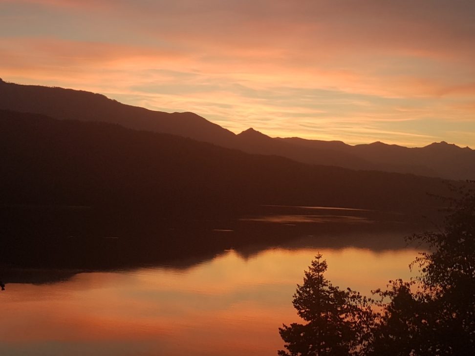 Sonnenuntergang über einem ruhigen See, umgeben von Bergen und buntem Himmel.